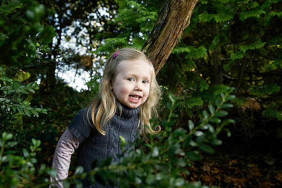 Little girl peeks out from behind a bush, smiling into the camera