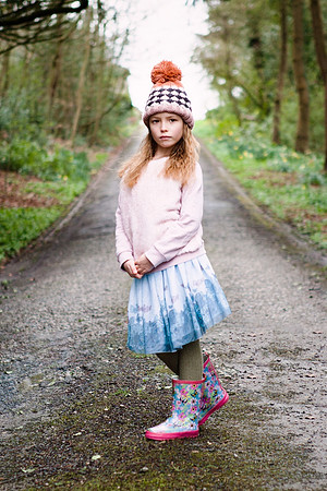 Little girl wears her party clothes with wellies and bobble hat, photographed in the woods