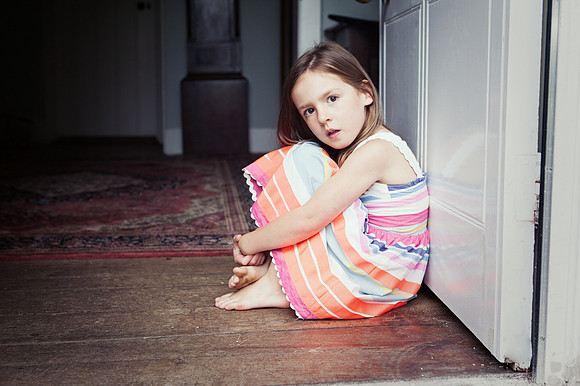A down eyed little girl sits in a doorway, just looking at the camera.  No smiles just a look.